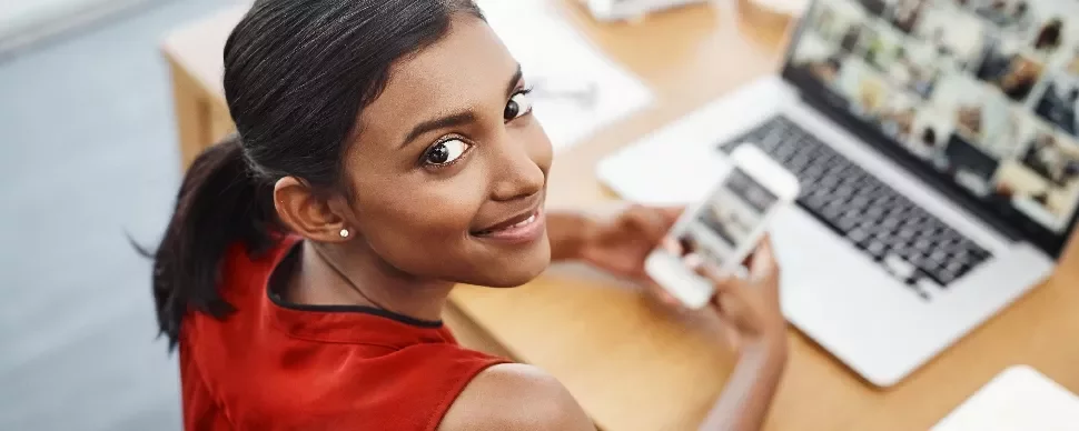 Woman in red top smiling, holding a smartphone while looking at a laptop on a wooden desk.