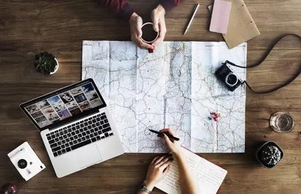 Map, laptop, camera, and notebooks on a wooden table with hands planning a trip.