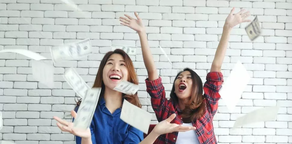 Two women happily throwing money in the air against a white brick wall.