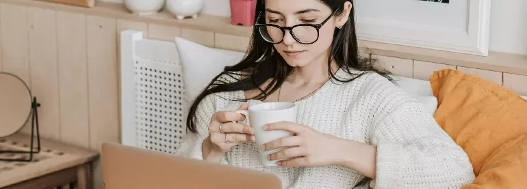 Woman in glasses reading on a laptop and holding a mug
