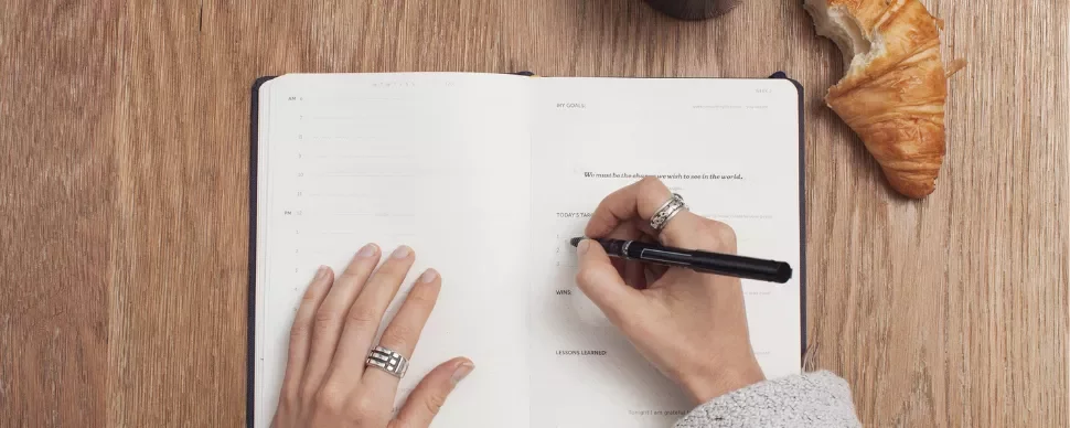 Person writing in a notebook with a pen, croissant on table.