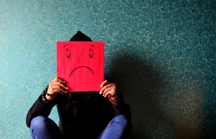 Person sitting against a textured wall holding a red paper with a sad face drawn on it.