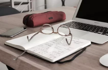 Open notebook with glasses on a desk near a laptop, phone, and pencil case.