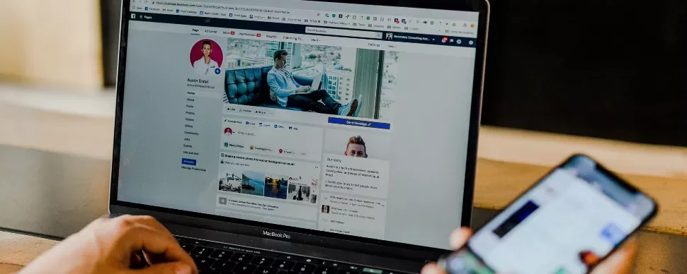 Person using a laptop, browsing a social media profile, while holding a smartphone.