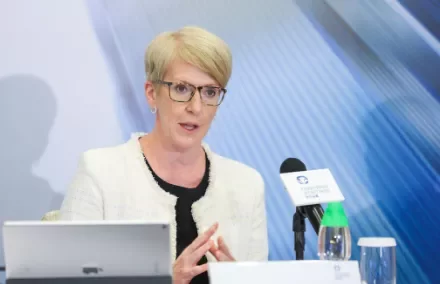 A woman with short blond hair and glasses speaks at a conference, seated with a tablet and microphone on the table.