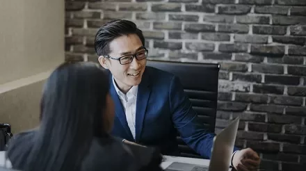 Two people in a meeting, one smiling at a laptop, in a modern office with a brick wall.